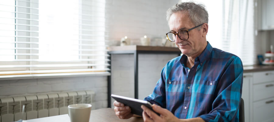 An older man with glasses wearing a blue plaid shirt sitting at a table looking at his tablet with a coffee cup nearby