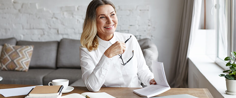 A woman in a white collared shirt smiling and holding her glasses and a book sitting at a table