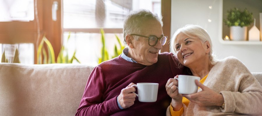 An older couple sitting on the couch holdings coffee cups smiling at each other