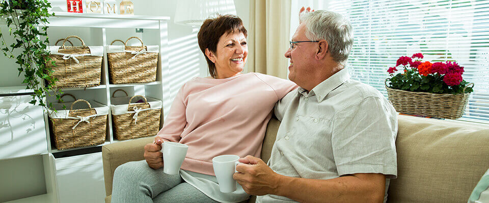 An older couple smiling at each other on the couch each holding a cup of coffee