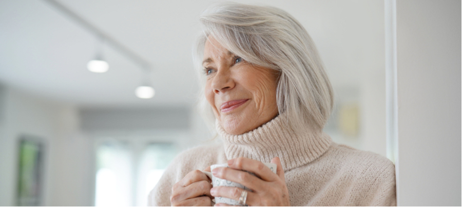 An older woman wearing a white turtleneck sweater holding a mug looking off in the distance