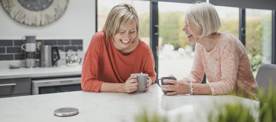 Two ladies laughing together at the kitchen counter holding coffee cups