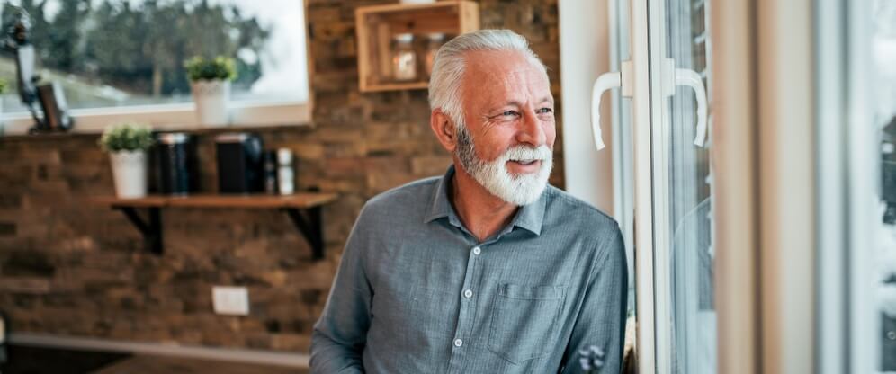 An older bearded man looking out the window in his home