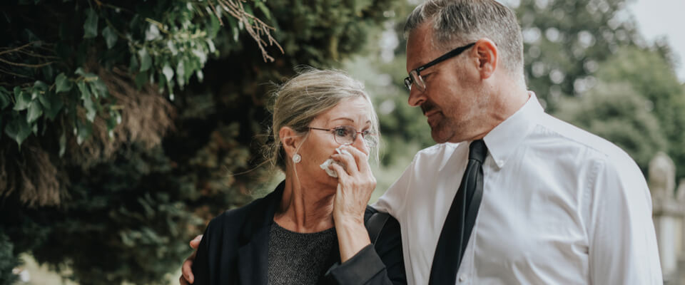 An older woman holding a tissue to her eyes while walking with a man wearing a white shirt and black tie