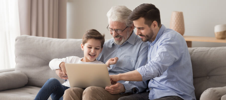 A grandfather holding the laptop with his son and grandson sitting on the couch