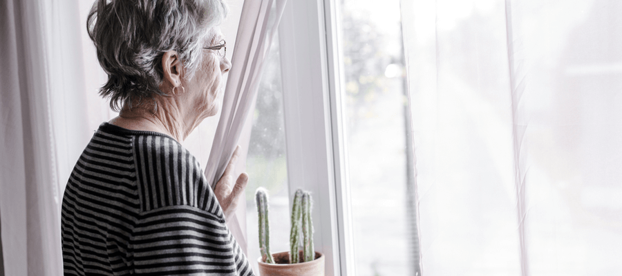 An older woman standing by the window holding the curtain aside to look out