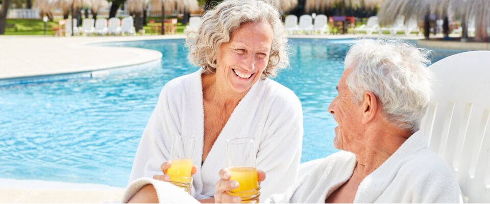 A couple wearing white robes sitting by a pool holding cups of orange juice