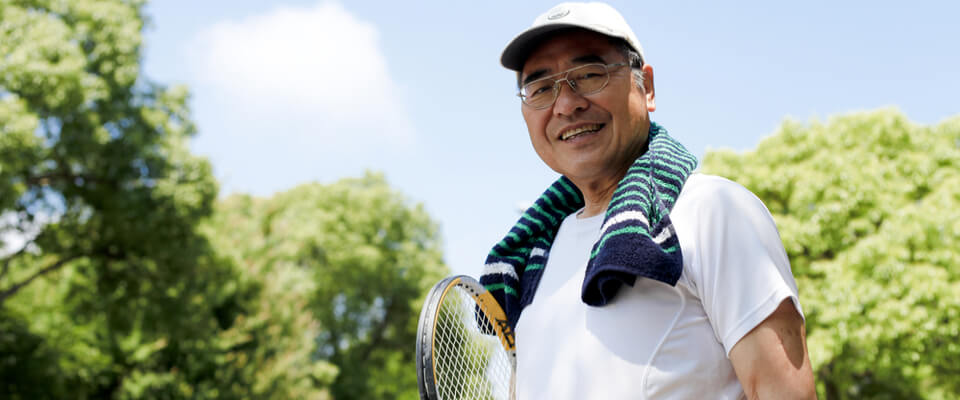An Asian man with glasses holding a tennis racket and tennis hat and towel smiling at the camera