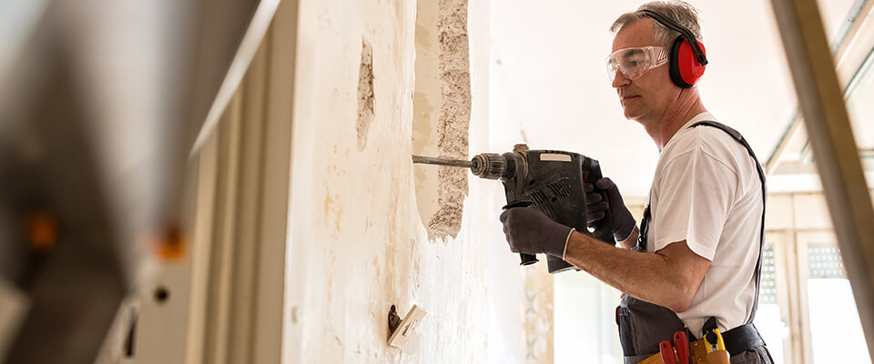 A man holding a drill working on a wall wearing safety gear