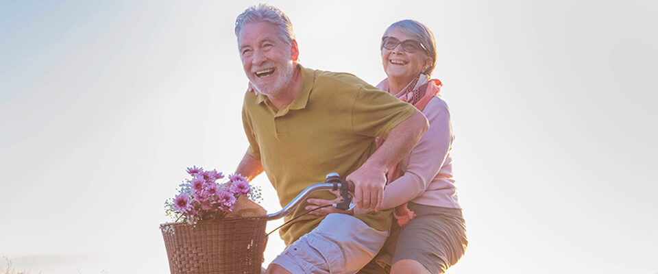 An older couple riding a bike together with a bouquet of flowers in the basket at the front of the bike