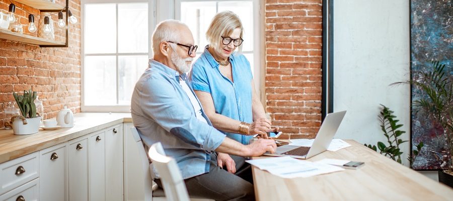 An older couple working at a kitchen counter holding mobile phone and working at laptop