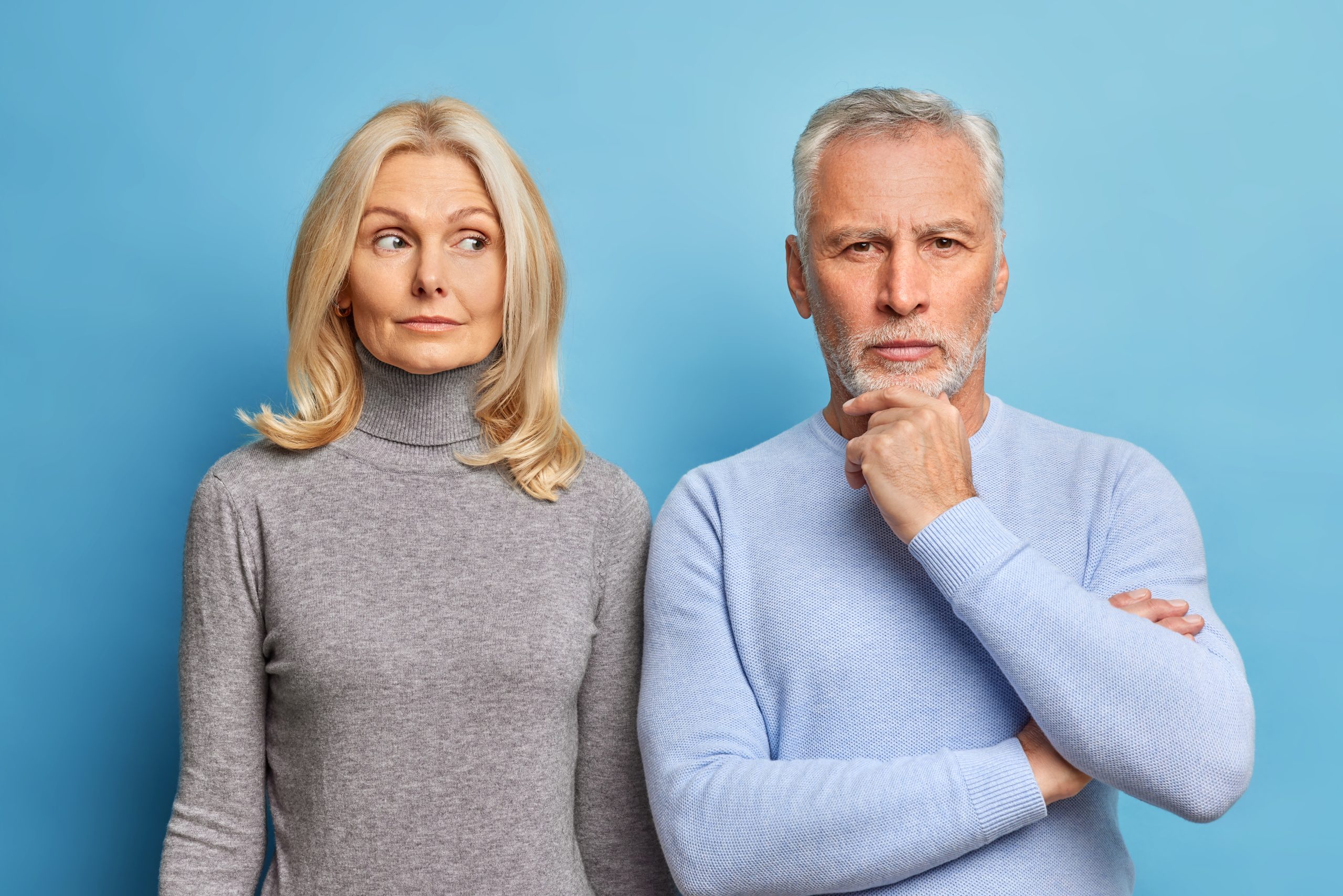 A serious older woman is standing next to an older man who appears to be thinking about something. Bright blue background, subjects clearly in view.