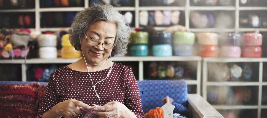 An older woman knitting and wearing headphones sitting on a couch